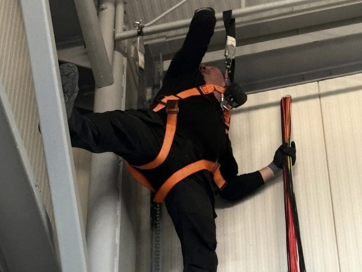 Installer in a fall-arrest harness pulling a bundle of cables under the roof of a hall